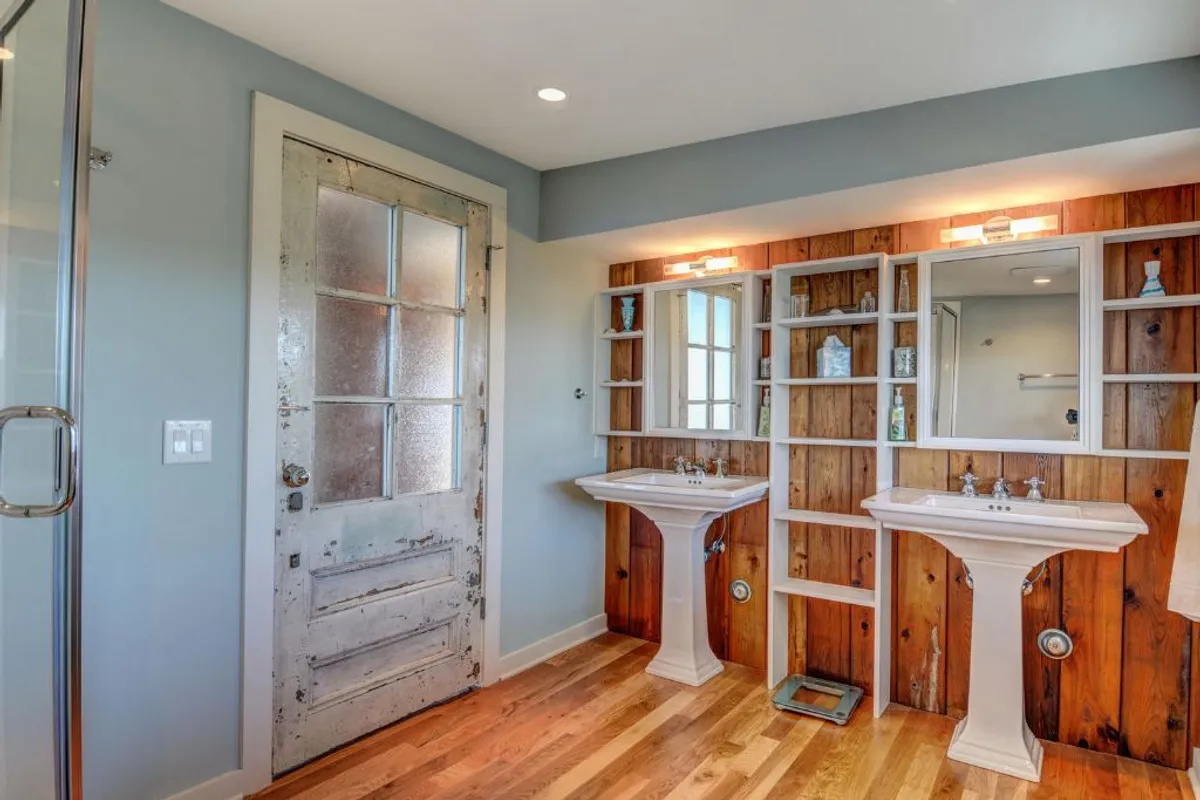 A cozy bathroom with dual sinks, a vintage salvaged door, wooden accent walls, and elegant mirrors. The design blends modern comfort with historical details, highlighting craftsmanship and a curated sense of style.