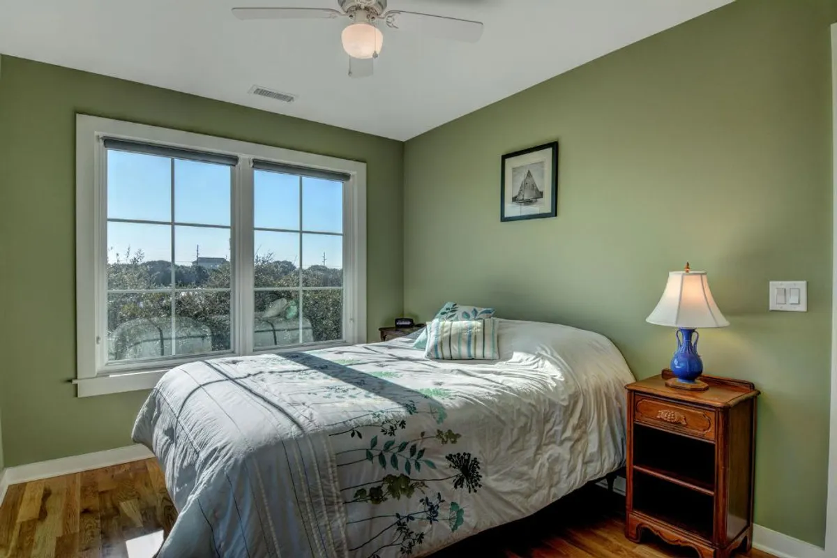 A serene master bedroom featuring a cozy bed with soft linens, a nightstand with a lamp, framed artwork, and large windows letting in natural light. The room exudes tranquility and invites restful sleep or quiet moments reading by the window.