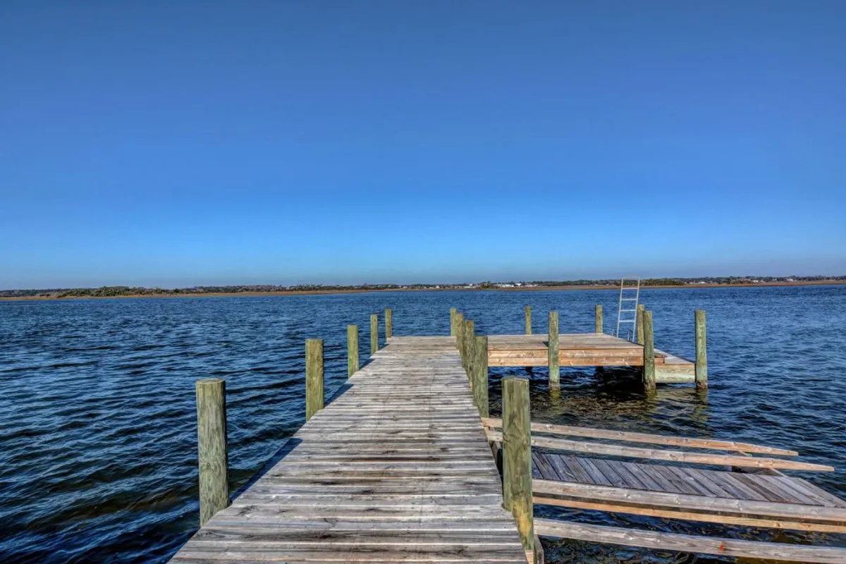 A serene wooden dock stretches out over calm water under a bright blue sky. The natural surroundings are peaceful, with gentle ripples on the water and the promise of sunset views. The scene hints at leisurely activities like fishing, sunbathing, or simply listening to the waves.