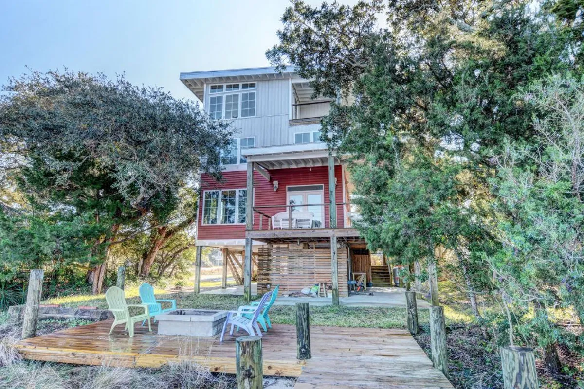 A cheerful exterior view of The Nancy Ann, featuring colorful Adirondack chairs arranged around a fire pit on a spacious deck. The two-story house is visible, with classic cottage architecture and inviting outdoor gathering spaces, all basked in natural sunlight.
