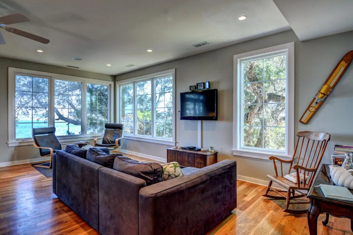 This image features a comfortable living area with large windows overlooking the water, a cozy seating arrangement including a wooden rocking chair, and a TV mounted on the wall. The view outside suggests a tranquil setting beneath expansive oaks, inviting relaxation and togetherness. The layout encourages conversations, games, and shared memories, all highlighted by the natural beauty visible through the windows.