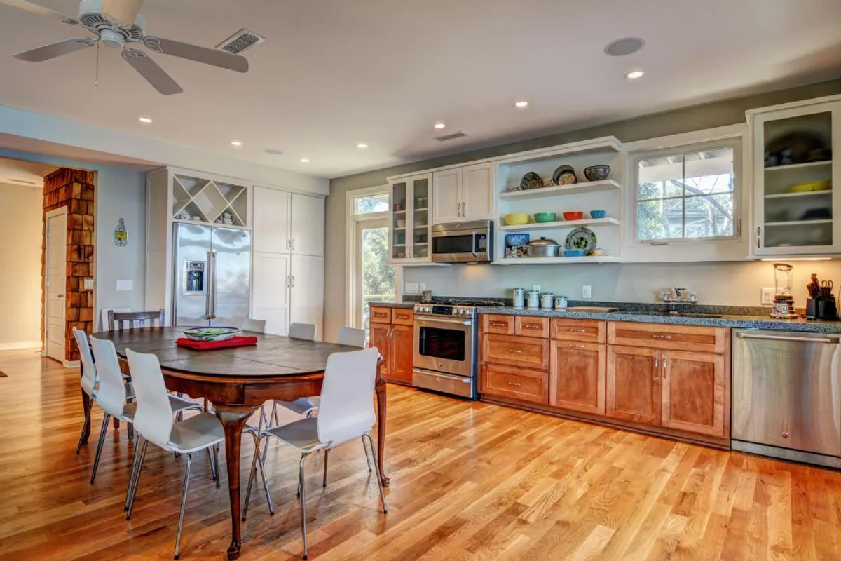The photo showcases a wooden dining table set in a bright, cheerful kitchen with stainless steel appliances and open shelving displaying curated dishware. Large windows flood the space with light, highlighting the warmth and character of the dining area.