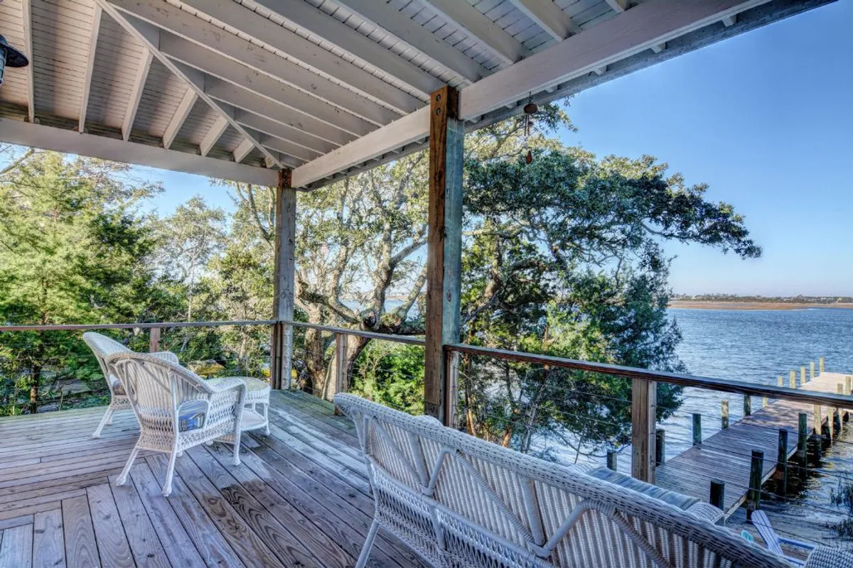 The image showcases a spacious wooden deck lined with inviting wicker chairs, overlooking tranquil waters and a distant dock framed by lush oak trees. The setting is serene, with sunrise rays gently illuminating the scene, promising peaceful moments spent relaxing, reading, or watching for local wildlife.