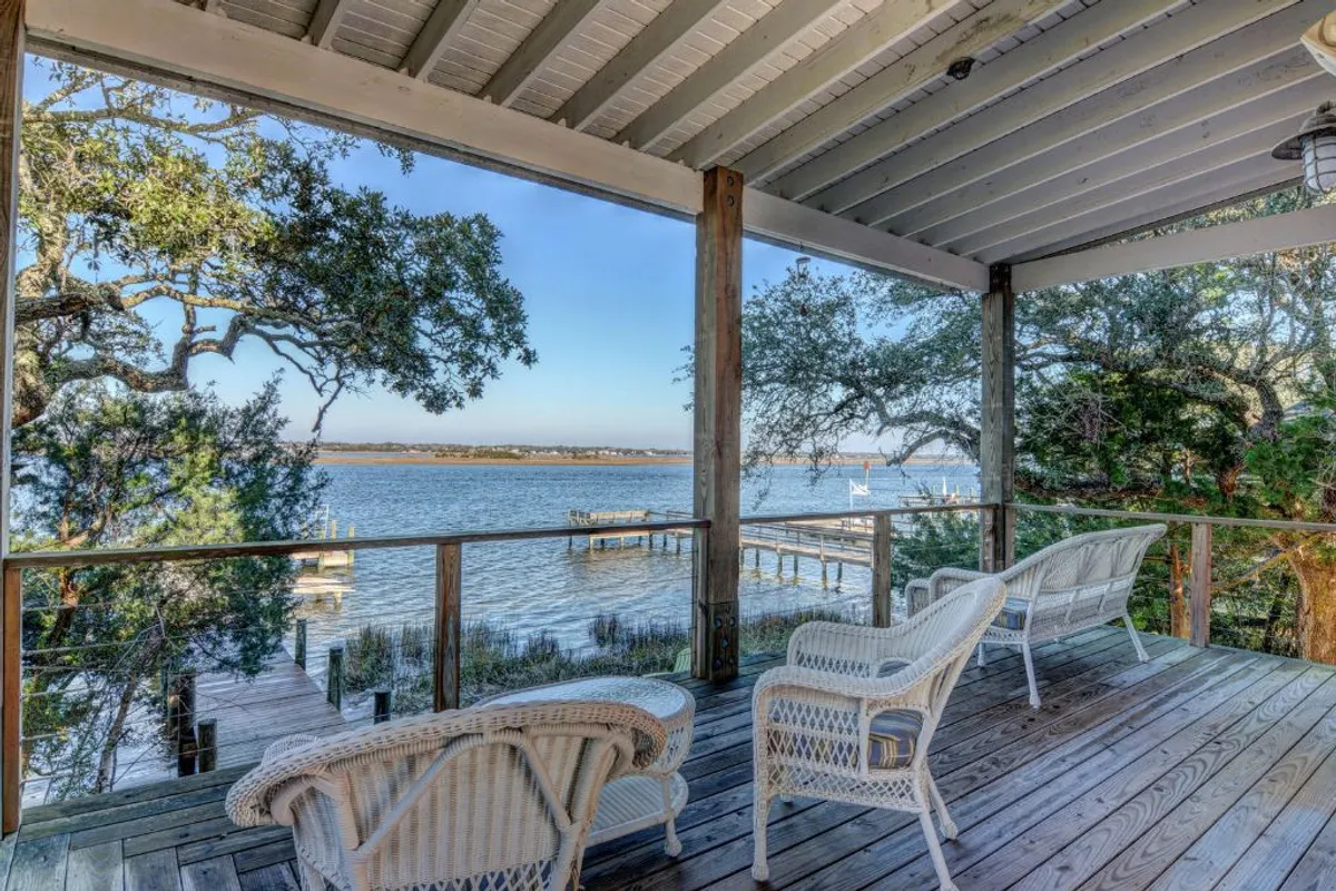 A serene outdoor deck is furnished with wicker chairs, providing a comfortable spot to relax and soak in views of the water and surrounding trees. The wooden deck seamlessly blends into the lush greenery, inviting guests to enjoy the tranquil outdoor atmosphere.