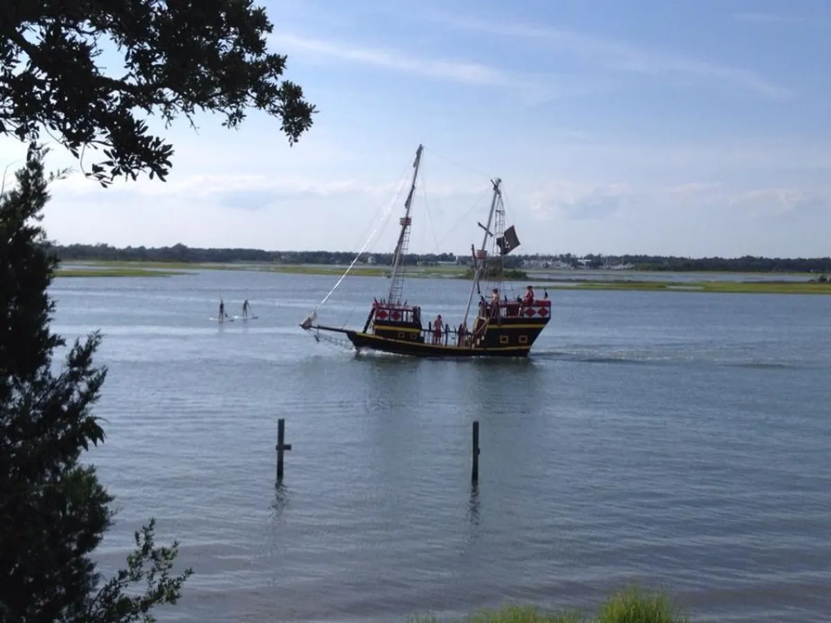 A vibrant waterfront scene featuring a pirate ship anchored nearby and several paddle boarders gliding over calm waters, with lush greenery framing the background. The mood is both adventurous and relaxed, evoking the spirit of exploration on the water.