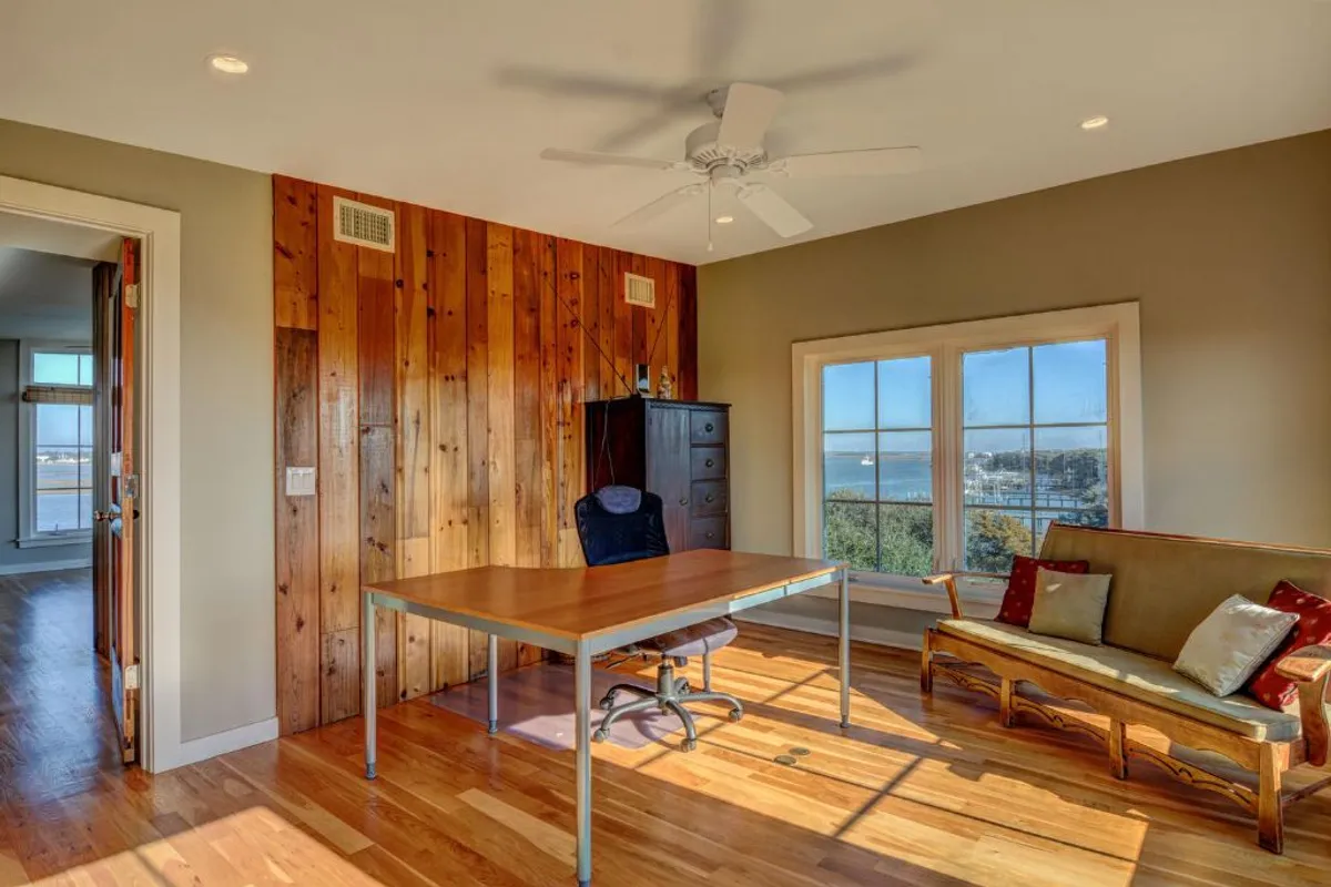 A calm office nook with a simple desk, comfortable seating, a wooden accent wall, and large windows offering inspiring views. The space is filled with natural light, providing an ideal environment for focused work or contemplation.
