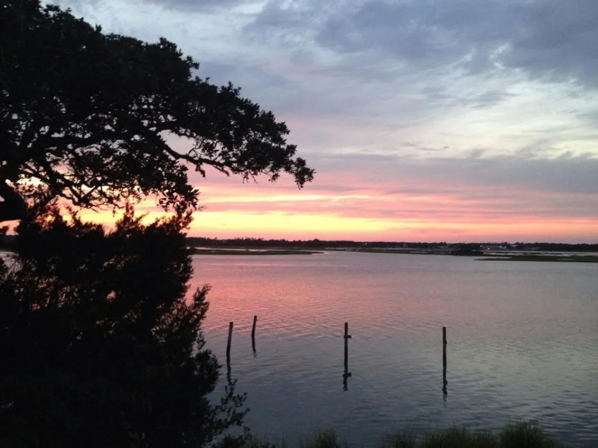 This scenic view shows a serene sunset sky reflected on calm water, with silhouetted trees and fishing posts, offering a sense of peacefulness and inspiration. The open expanse of nature encourages relaxation and creative thought, reinforcing the property's tranquil environment.