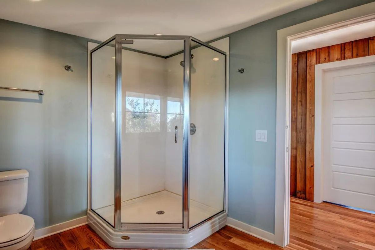 A bright, modern bathroom with a glass shower enclosure, wooden floors, and light blue walls. Morning sunlight streams in, highlighting the clean, serene design ideal for relaxation and self-care.