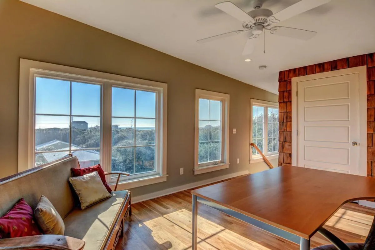 A relaxed living space with large windows, a wooden table, and comfortable seating. The atmosphere is cozy and relaxed, with natural light streaming in and a sense of connection to the water and surrounding landscape.